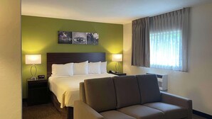 Ein Hotelzimmer mit einem Bett, zwei Nachttischen mit Lampen, einem Sofa, einem Fenster mit Vorhängen und einer Wand mit drei gerahmten Bildern.