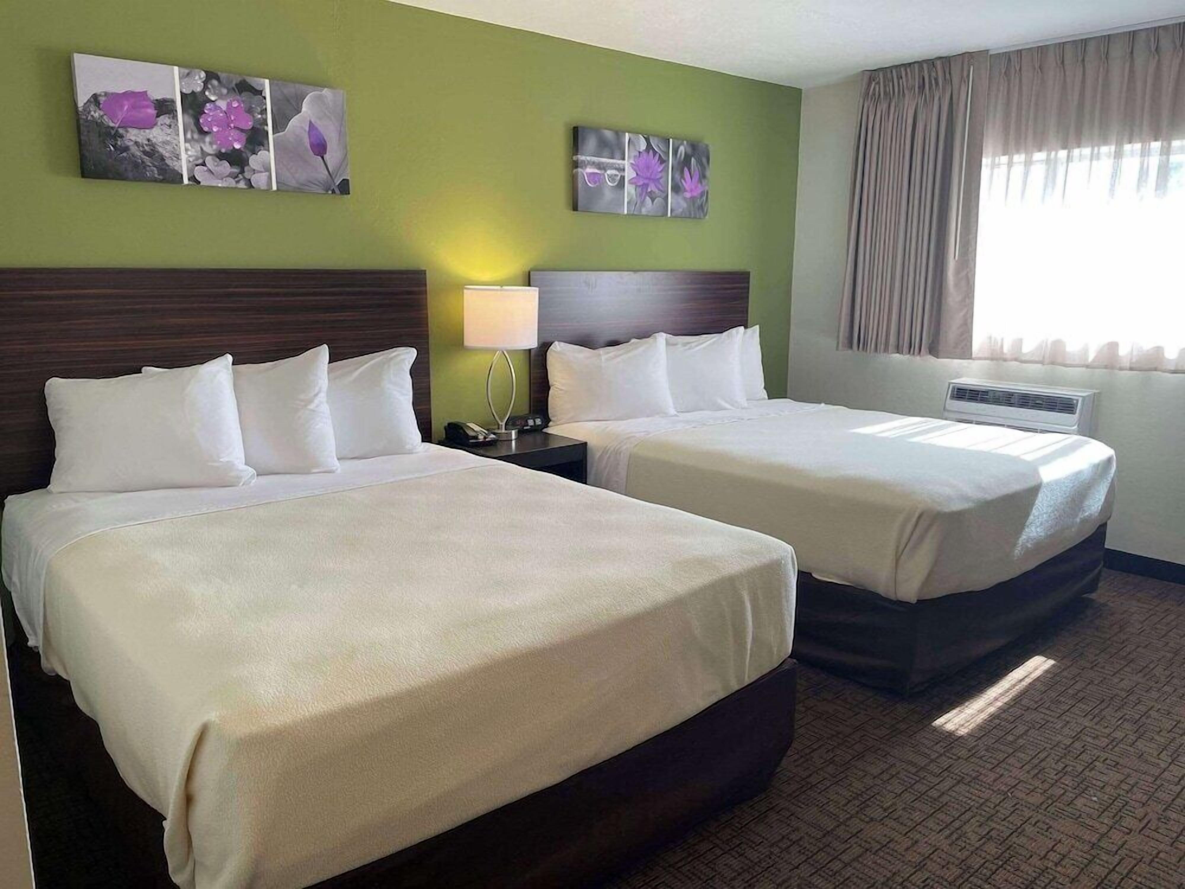 A hotel room with two beds, a nightstand with a lamp, a wall-mounted air conditioner, and artwork above the beds.