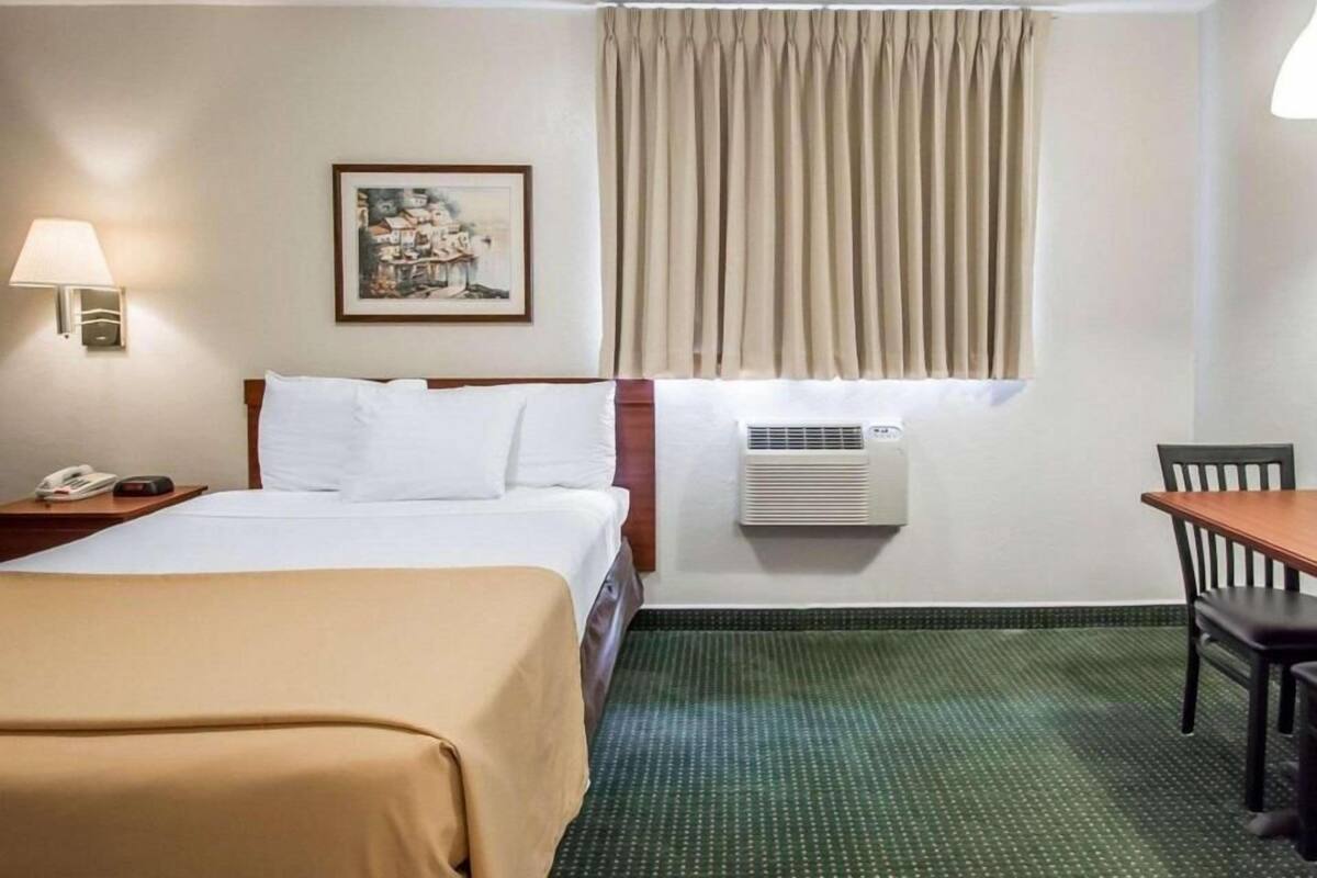 Een hotelkamer met een bed, nachtkastje, lamp, airconditioning en een klein tafeltje met stoel.