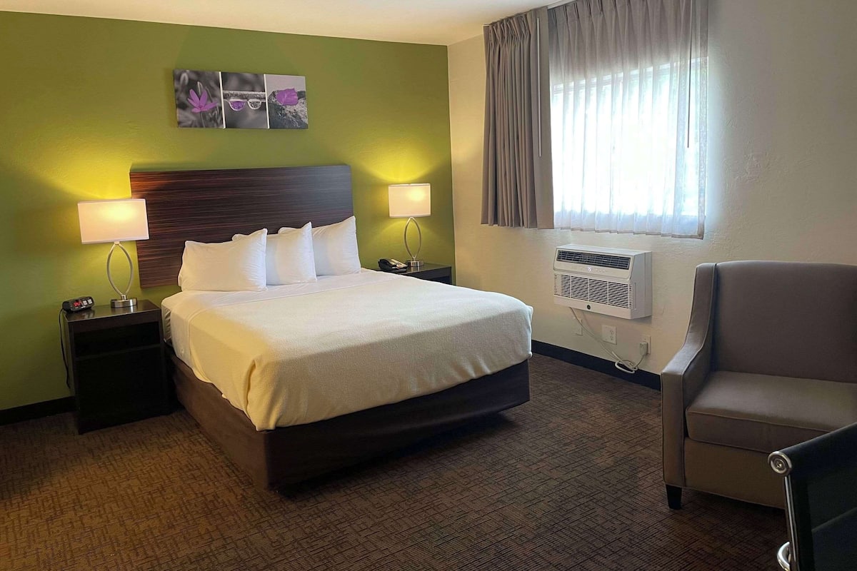 Een hotelkamer met een bed, twee nachtkastjes met lampen, een stoel, een raam met gordijnen en een airconditioning.