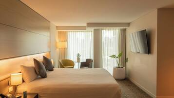 A hotel room with a bed, bedside table with a lamp, a chair, a small table with a plant, and a TV mounted on the wall.