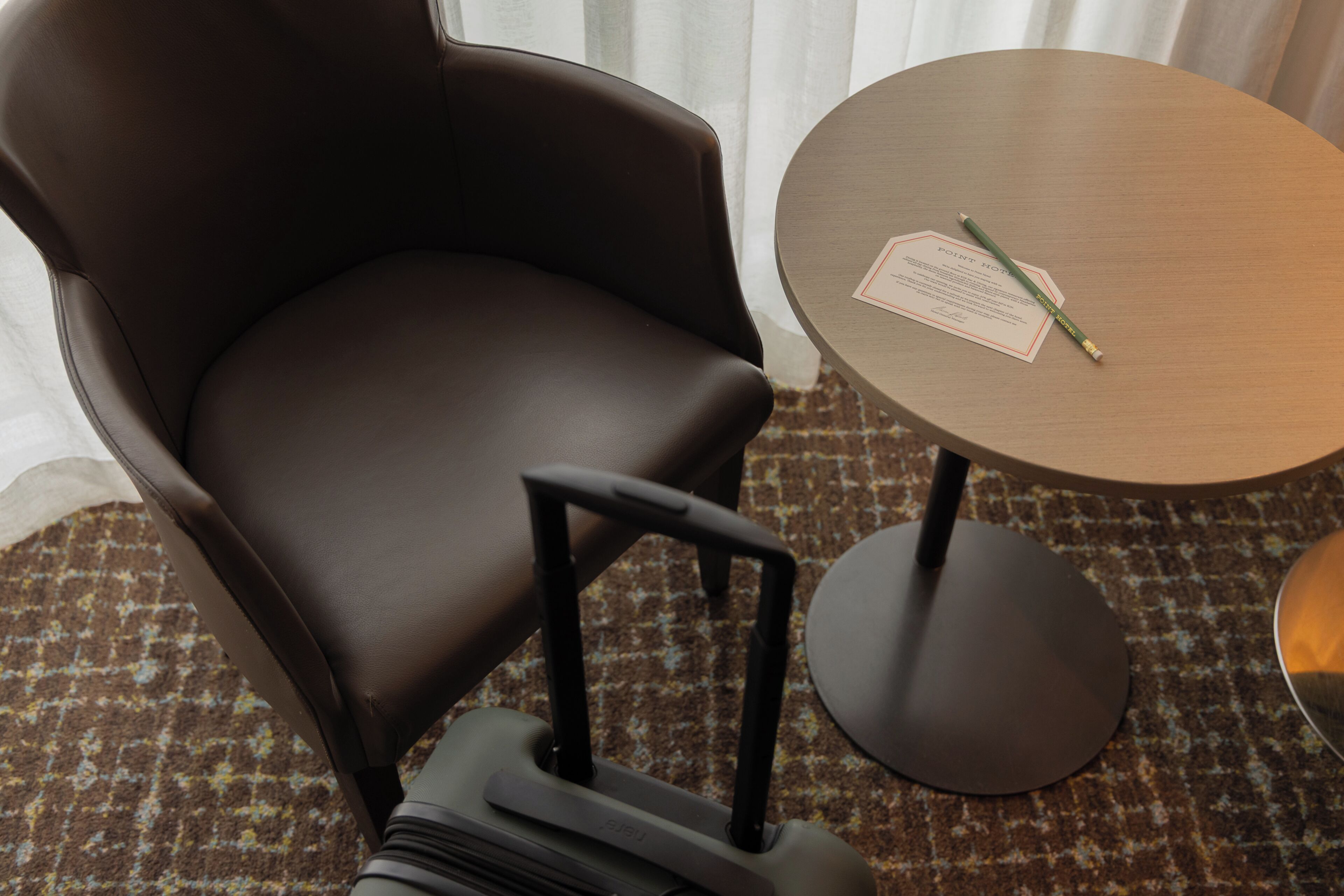 A brown leather chair with a footrest and wheels, a small round table with a document and pencil, and a patterned carpet.