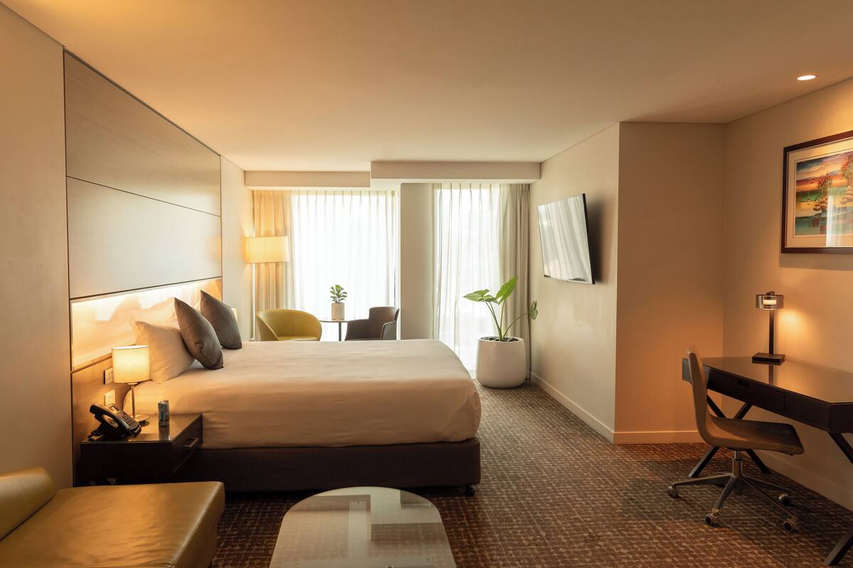 A hotel room with a bed, desk, chair, lamp, and a TV on the wall.