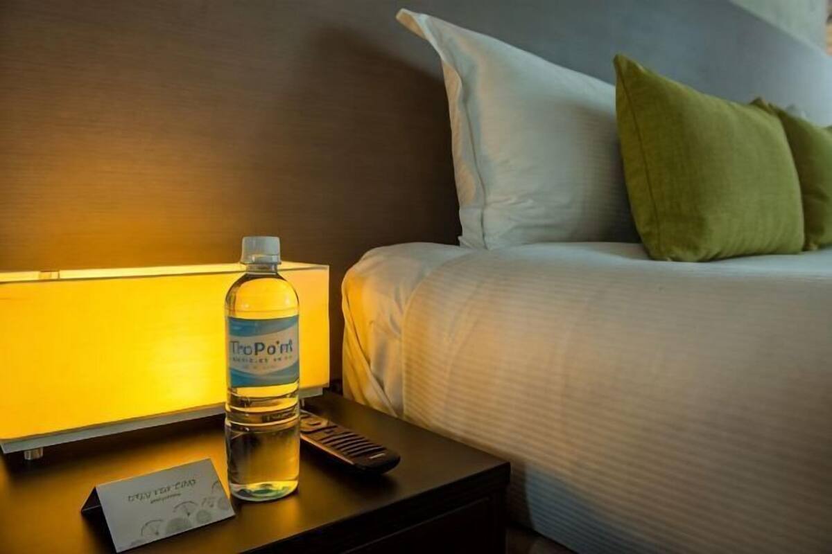 A hotel room with a bed, a nightstand with a bottle of water, a remote control, and a card on the nightstand.
