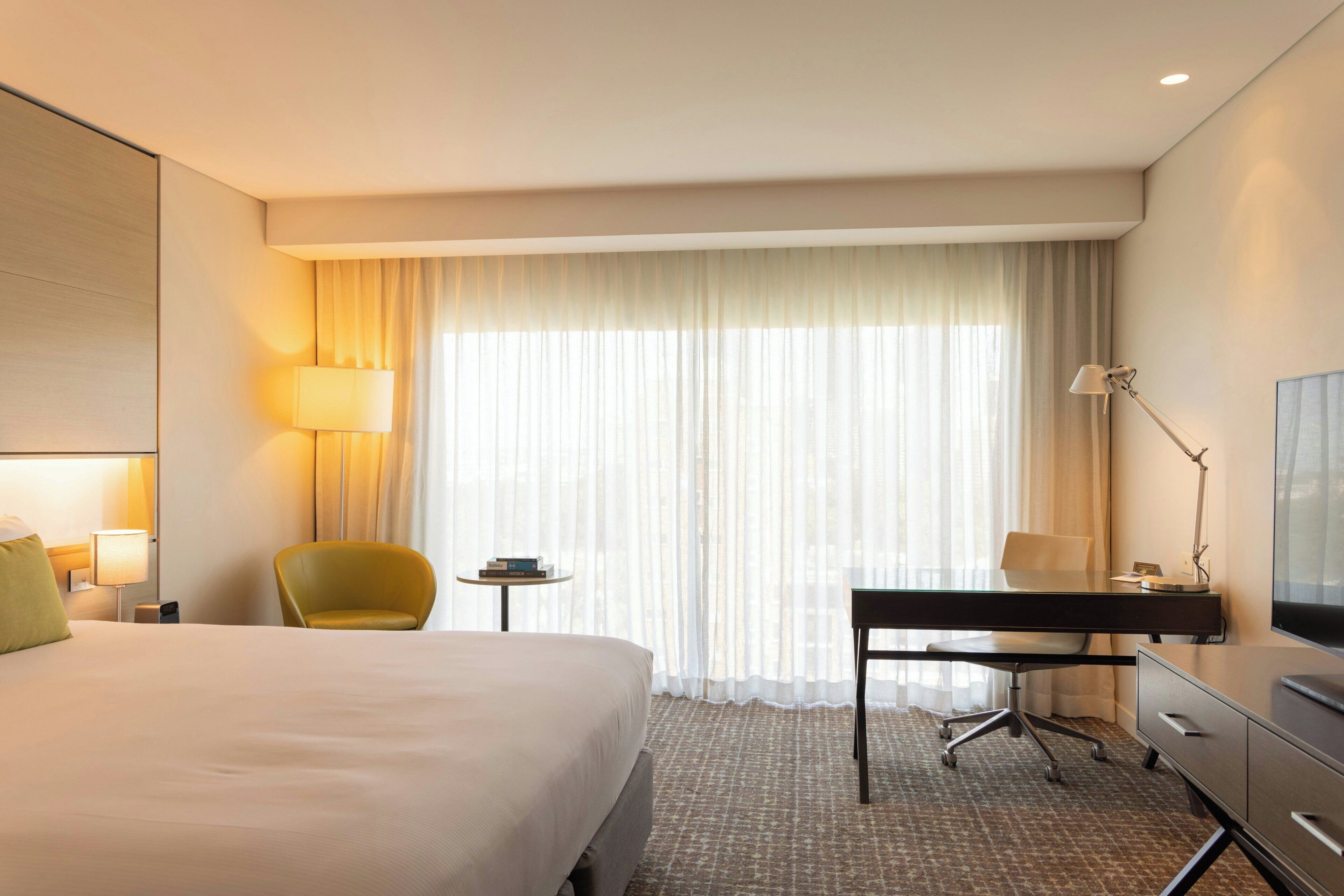 Una habitación de hotel con cama, escritorio, silla, lámpara, televisor y ventana con cortinas.