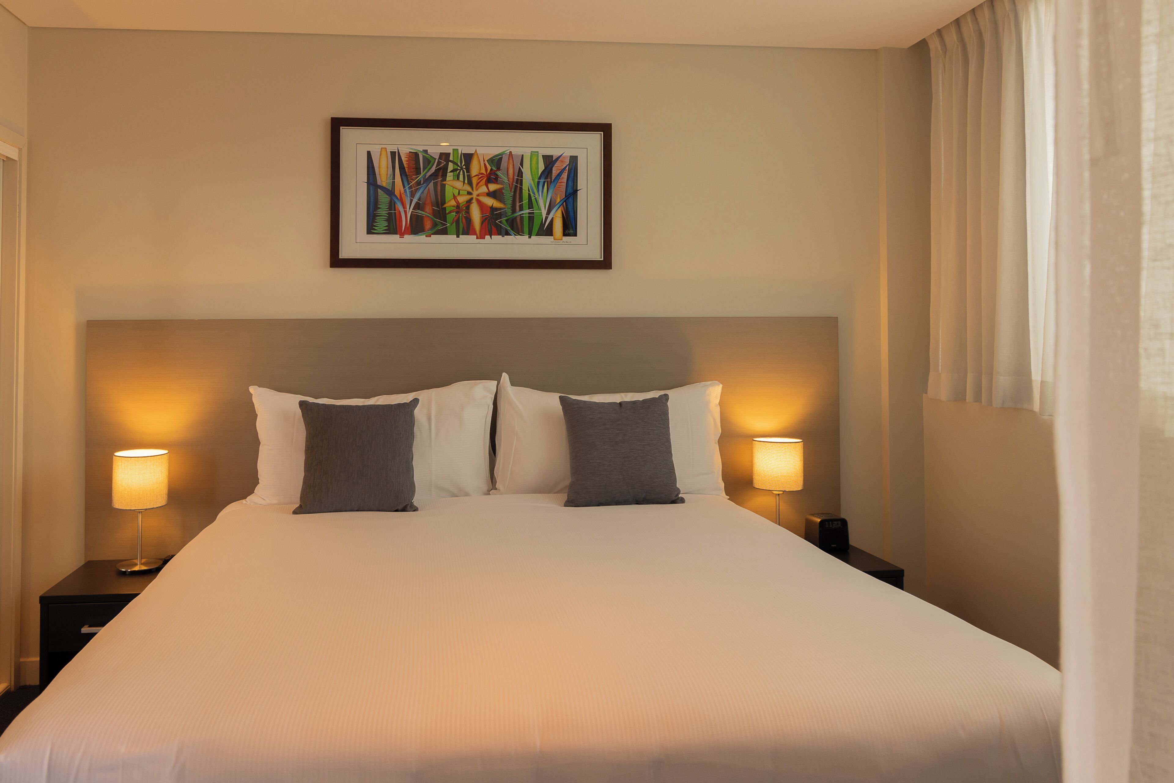 A hotel room with a large bed, two bedside lamps, a framed abstract painting, and a window with curtains.