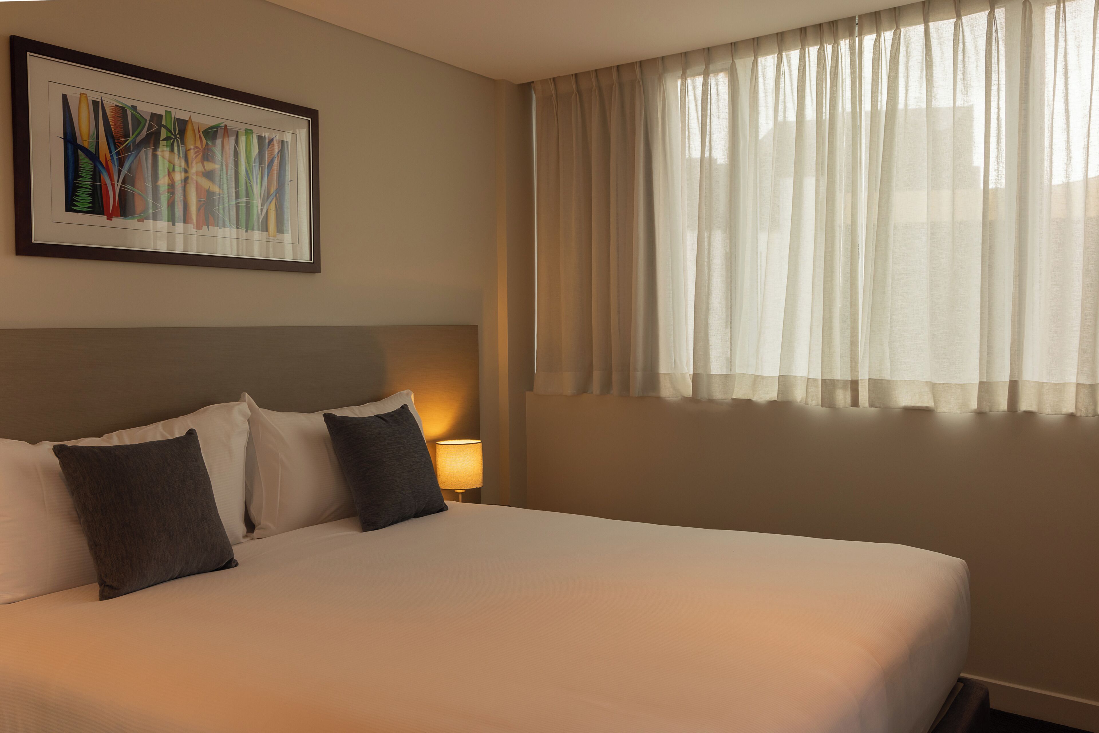A hotel room with a large bed, a framed artwork above the bed, a bedside lamp, and sheer curtains on the window.