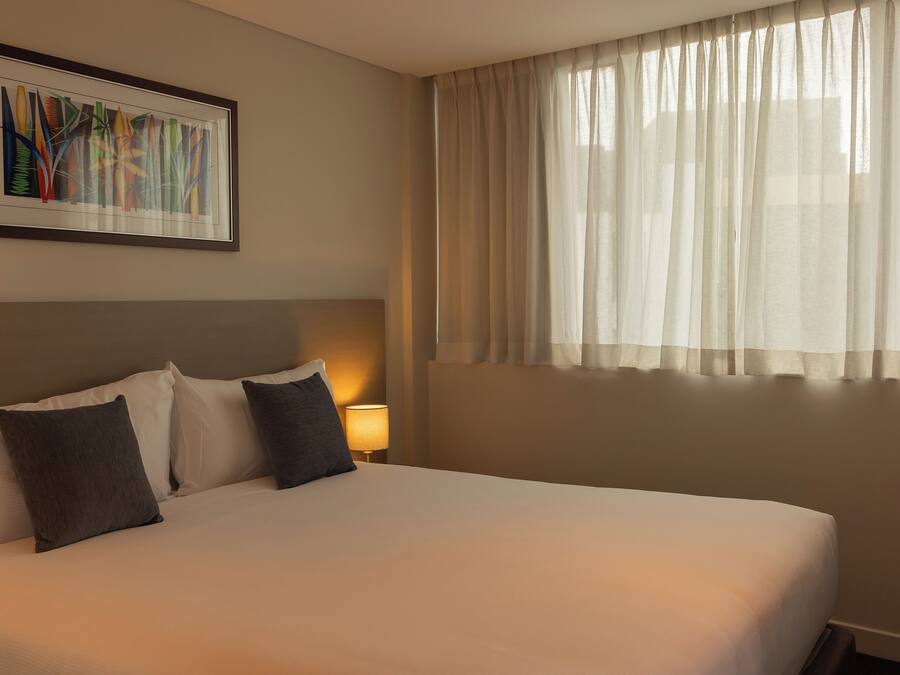 Een hotelkamer met een groot bed, een ingelijst kunstwerk erboven, een bedlampje en dunne gordijnen voor het raam.