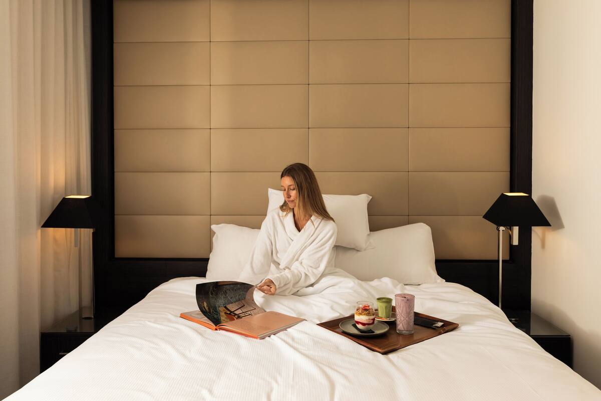 A hotel room with a bed, a bedside table, a lamp, and a tray with a book, a glass, and a dessert.