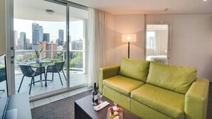 A modern living room with a green sofa, a dining table with chairs, a coffee table with a bottle and glasses, and a city view through large windows. - Point Hotel (Kangaroo Point)
