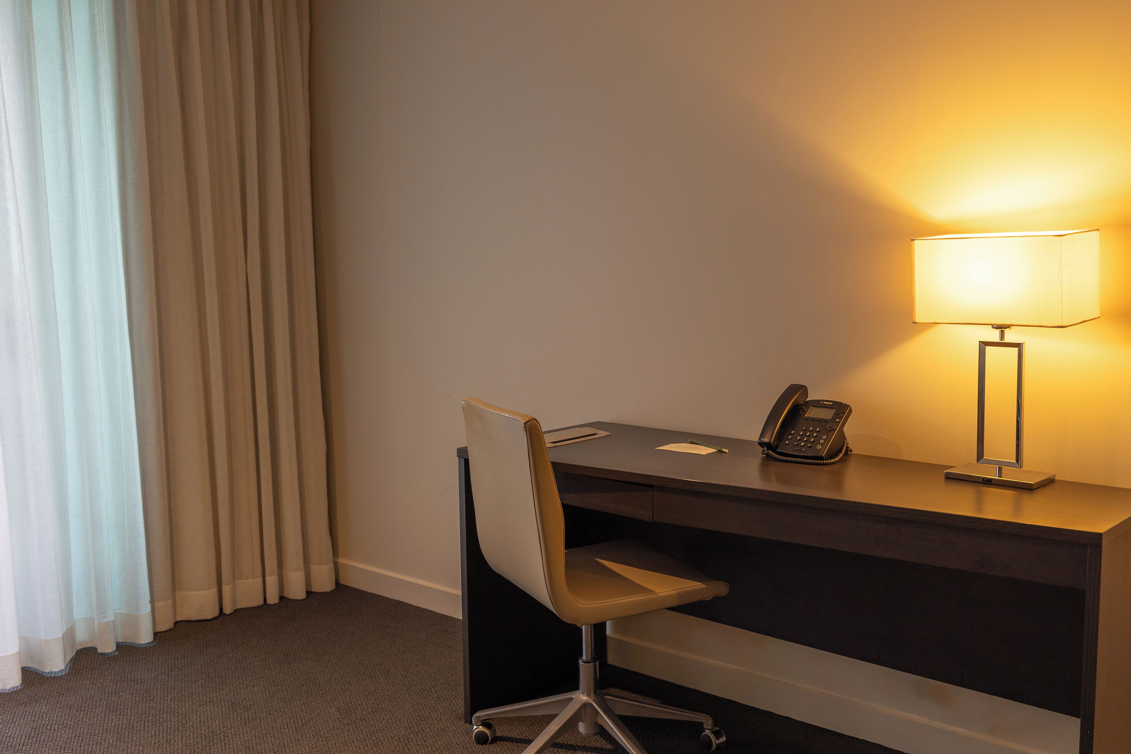 A hotel room with a desk, chair, lamp, phone, and curtains.