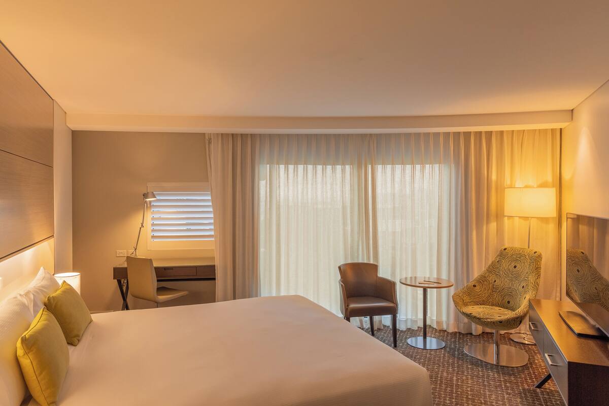 A hotel room with a bed, a desk, a chair, a small table, and a lamp.
