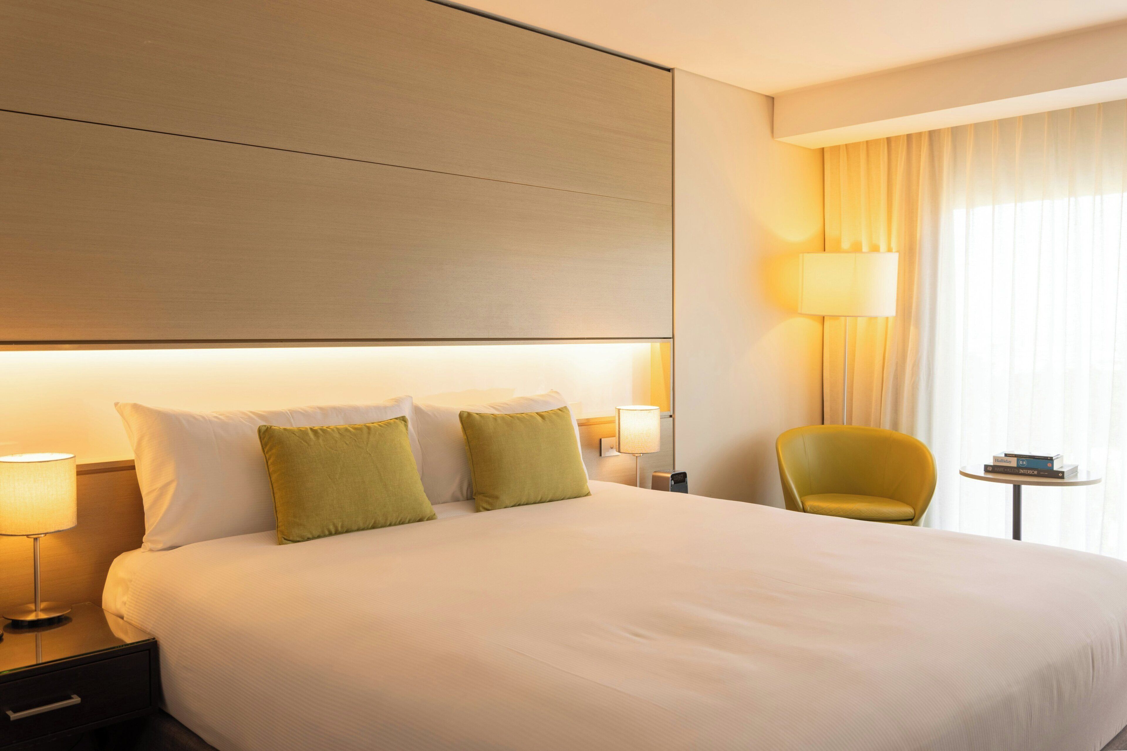 A hotel room with a large bed, bedside lamps, a small table, a chair, and a window with curtains.