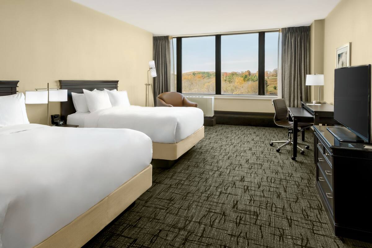 A hotel room with two beds, a desk, a chair, a TV, and a large window offering a view of the outdoors.