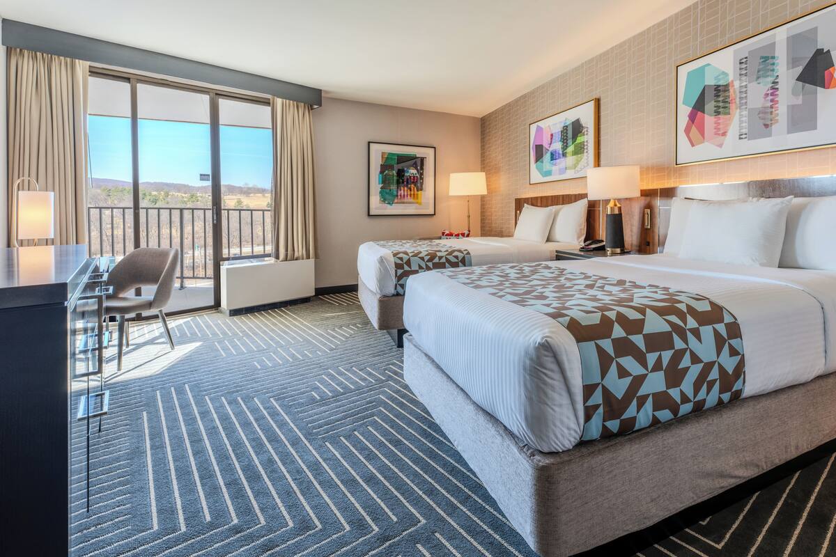 A hotel room with two beds, a desk, and a balcony with a view.