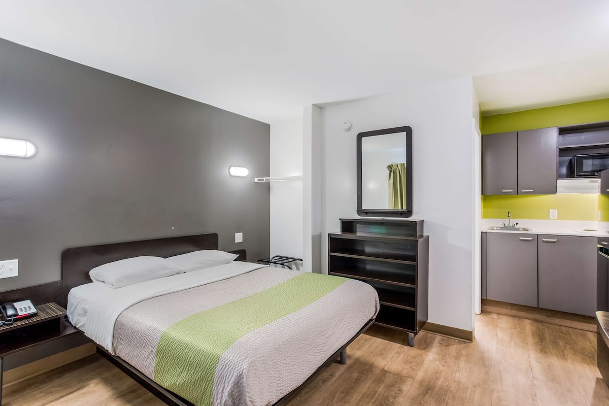 A compact hotel room with a bed, bedside table, and a small kitchenette area.