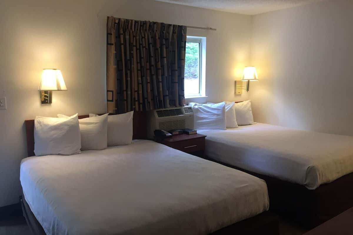 A hotel room with two beds, each with white linens and pillows, a bedside table with a lamp, and a window with curtains.