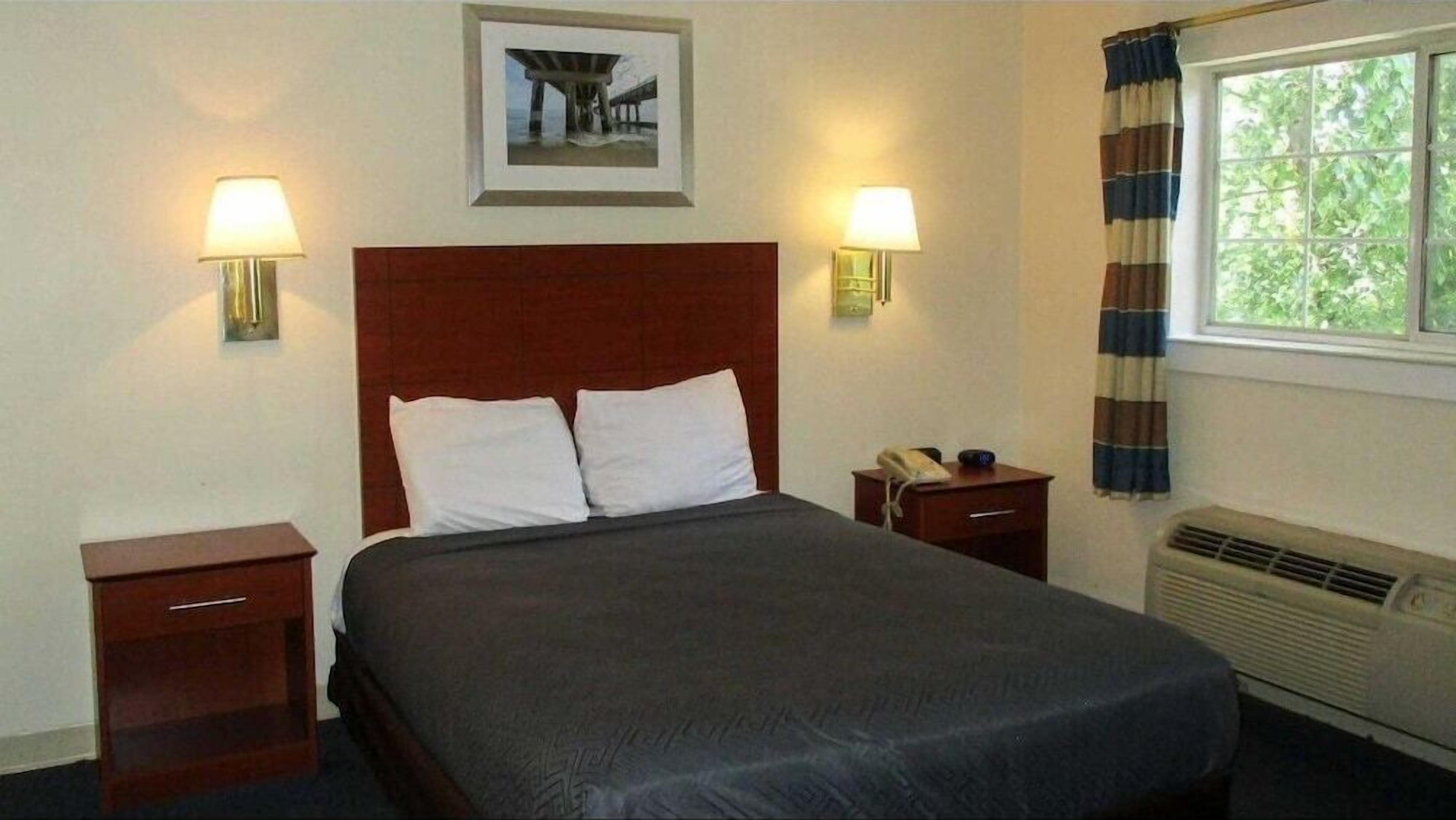Quarto de hotel com uma cama grande, duas mesas de cabeceira, um criado-mudo, um quadro na parede, uma janela com cortinas e um aquecedor fixado na parede.