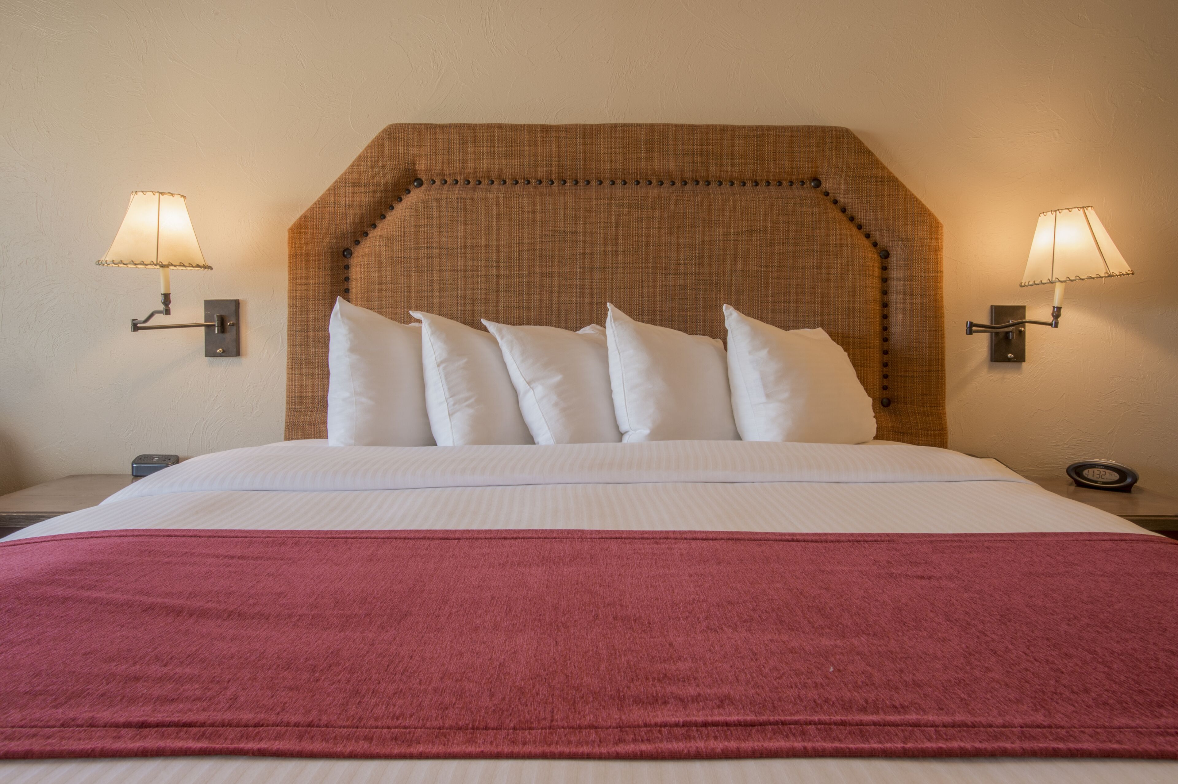 A neatly made bed with a red blanket, a headboard with a textured pattern, and two wall-mounted lamps on either side.