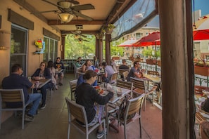 Enjoy al fresco dining in a shaded outdoor space with tables and chairs, surrounded by lush greenery and urban views.