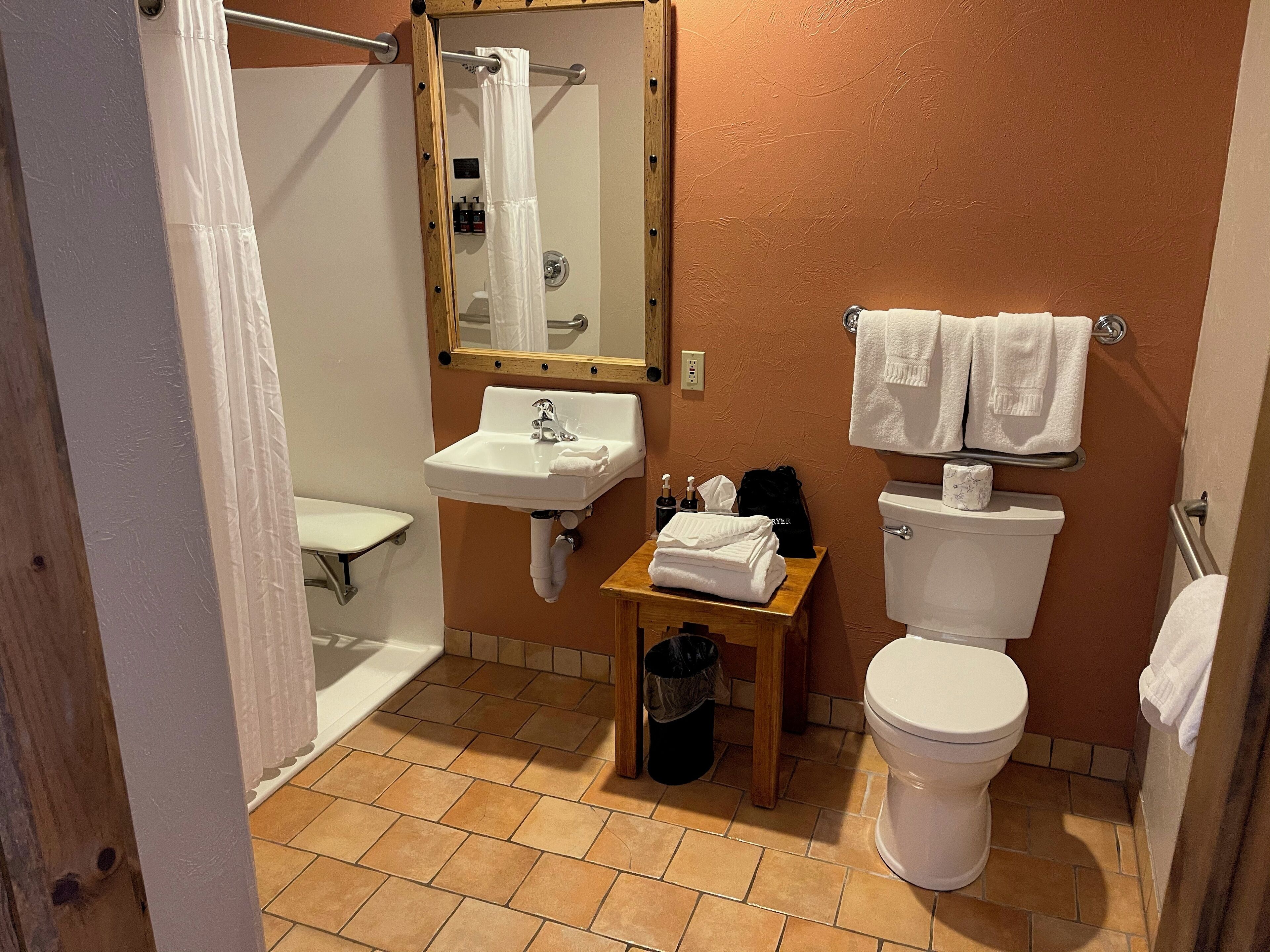 Un baño con lavamanos, inodoro y ducha. Hay una mesita con toallas y artículos de aseo.