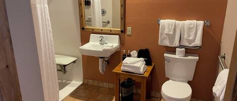 Un baño con lavamanos, inodoro y ducha. Hay una mesita con toallas y artículos de aseo.