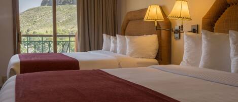 A hotel room with two beds, a balcony with a view of a mountainous landscape, and two wall-mounted lamps.