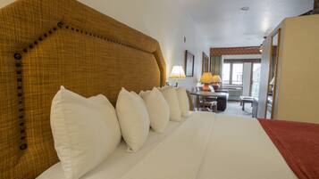 Habitación de hotel con una cama grande, varios almohadones, cabecera de madera, escritorio con lámpara y vista al exterior.