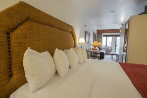 A hotel room with a large bed, multiple pillows, a wooden headboard, a desk with a lamp, and a view of the outside.