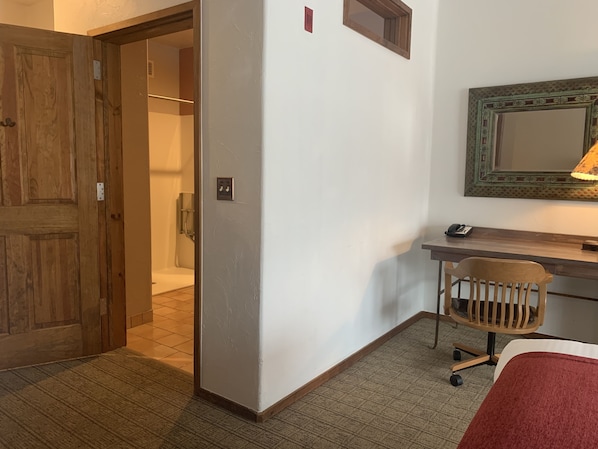 Habitación de hotel con puerta de madera, ducha, escritorio con silla y espejo.