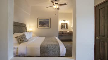 A hotel room with a bed, a ceiling fan, a bathroom with a mirror, and a mountain landscape painting on the wall.