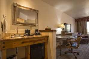 A hotel room with a wooden desk, a chair, a small refrigerator, a sink with a mirror, a television, and a lamp.