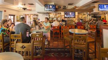 A bar with wooden chairs and tables, multiple TVs on the wall, and a variety of bottles on shelves.