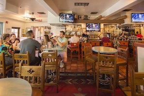 A bar with wooden chairs and tables, multiple TVs on the wall, and a variety of bottles on shelves.