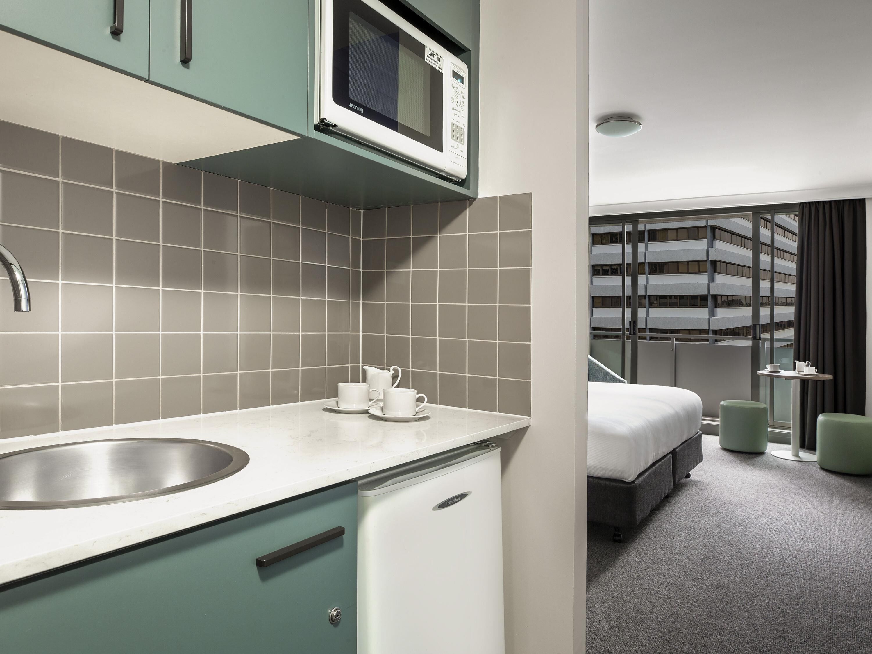 A modern hotel room with a kitchenette, a bed, and a view of a building.