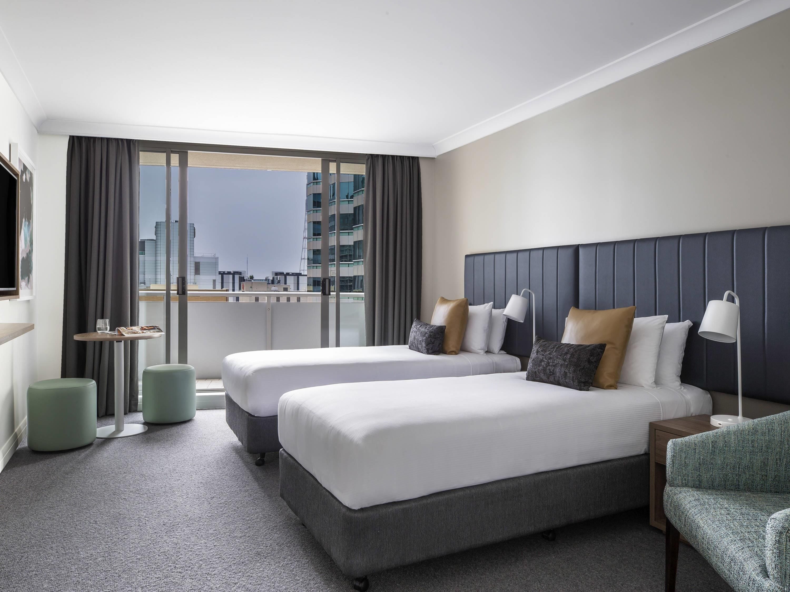 A hotel room with two beds, a small table, a chair, and a view of the city.