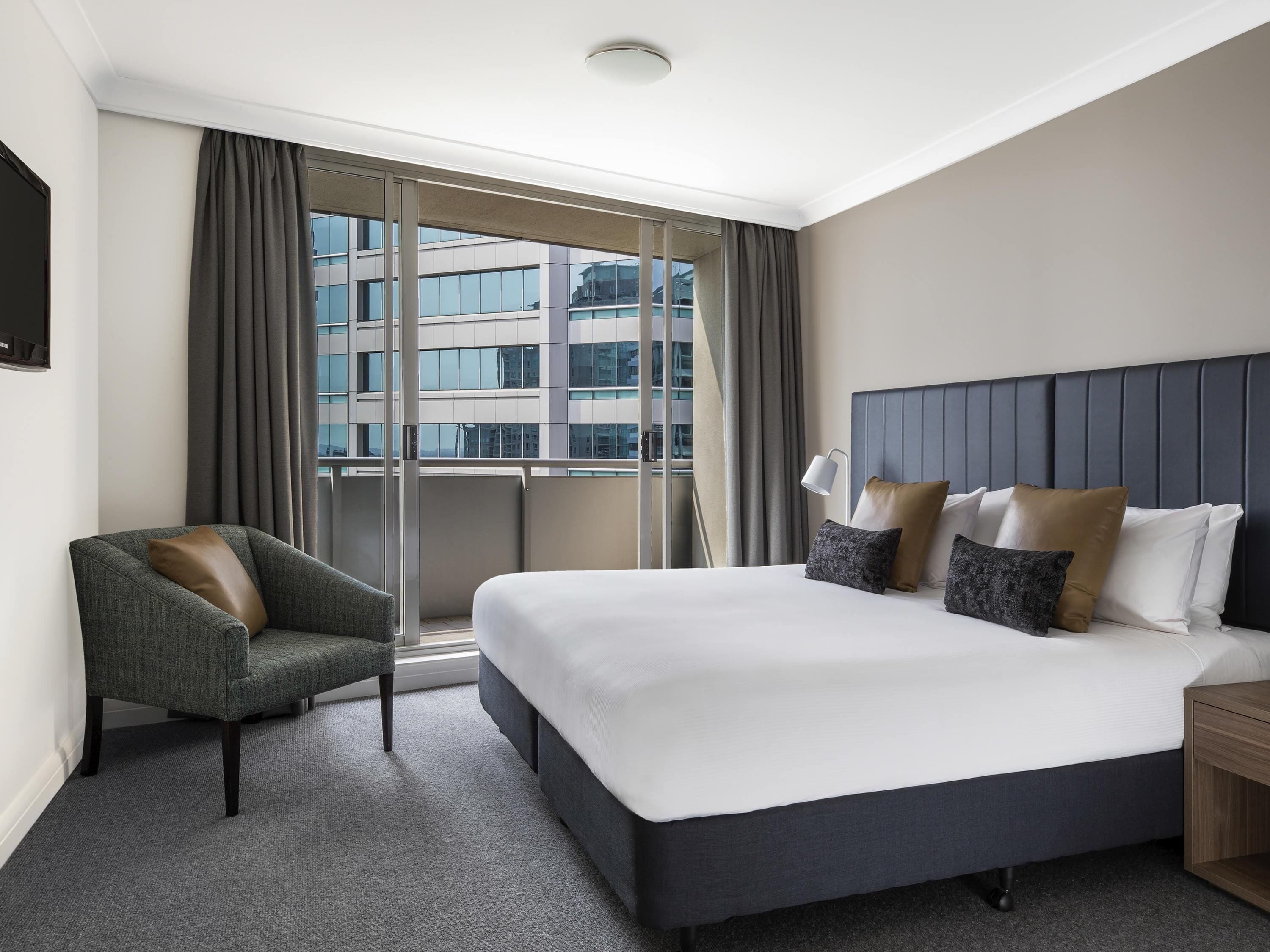 Ein Hotelzimmer mit einem großen Bett, einem Sessel, einem Nachttisch und Blick auf ein Gebäude durch das Fenster.