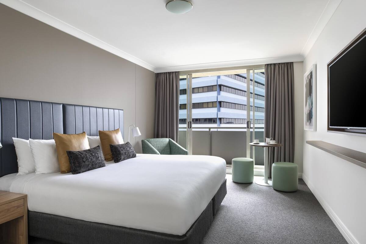 A hotel room with a large bed, a flat-screen TV, a small table with a cup and a pitcher, a chair, and a view of a building through the window.