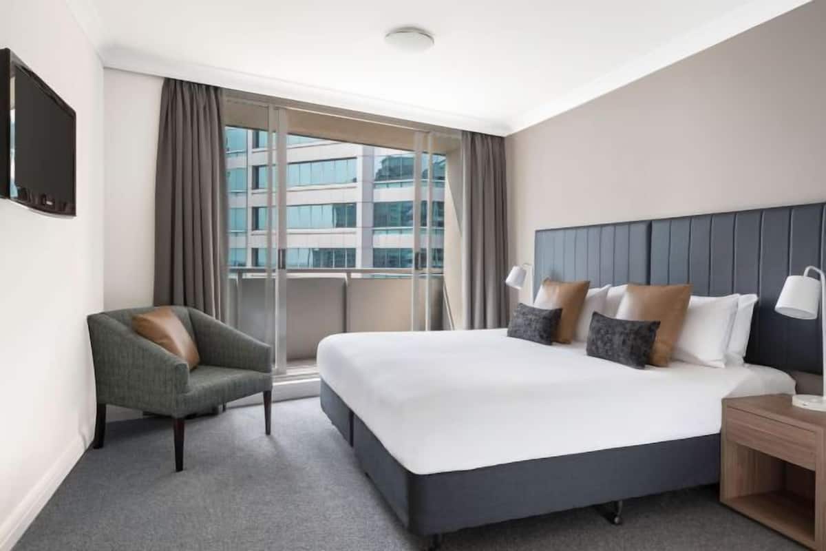 A hotel room with a large bed, a chair, a nightstand, a TV, and a window with curtains.
