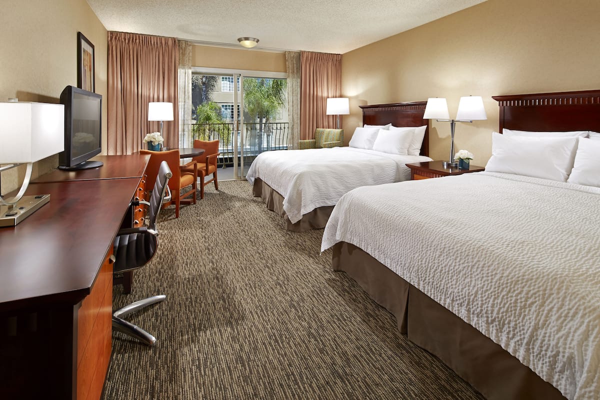 A hotel room with two beds, a desk with a TV, a chair, and a balcony with curtains.