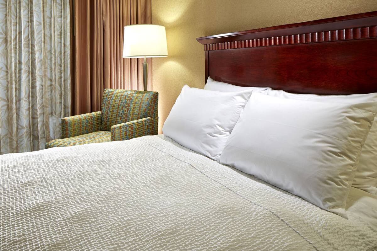 A hotel room with a large bed, a wooden headboard, a patterned armchair, and a lamp.