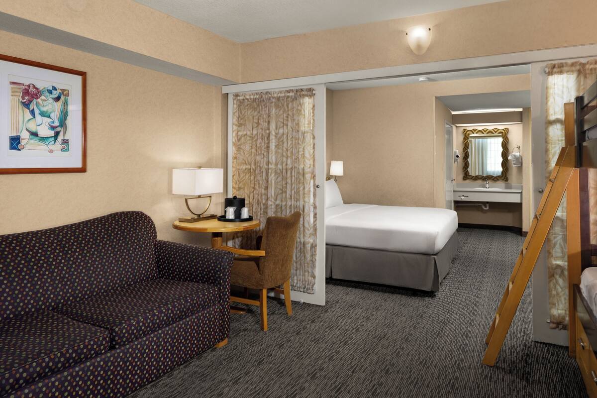 A hotel room with a sofa, a small table with a lamp, a bed, a mirror, and a wooden ladder.