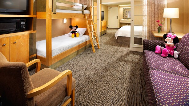 A hotel room with a bunk bed, a television, a desk with a lamp, a chair, and a couch with plush toys. - Anaheim Portofino Inn and Suites (Anaheim)