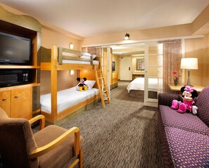 Una habitación de hotel con literas, televisión, escritorio con lámpara, silla y sofá con juguetes de peluche.