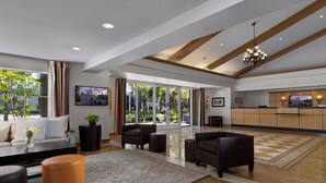 A spacious living area with a large rug, sofas, and a coffee table. There are large windows with curtains, a chandelier, and a kitchen area with a TV. - Anaheim Portofino Inn and Suites (Anaheim)