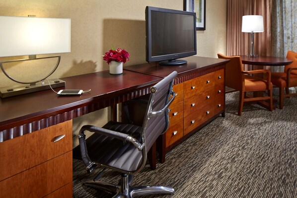 Una habitación de hotel con un escritorio de madera, un televisor de pantalla plana, una computadora portátil, un teléfono inteligente, una silla, una lámpara y un florero con flores.