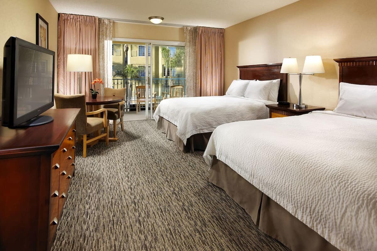 A hotel room with two beds, a desk with a chair, a television, and a balcony with curtains.
