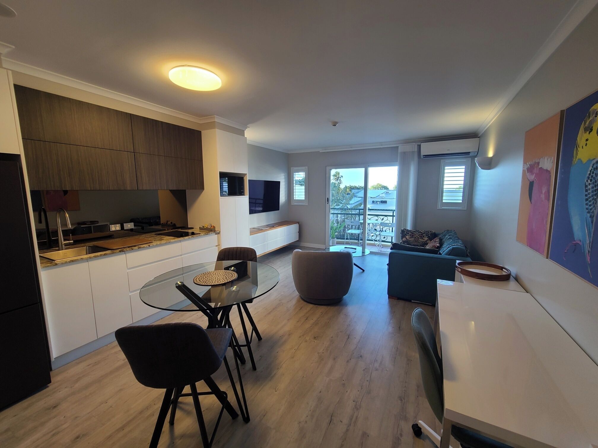 A modern kitchen with a dining area, a TV mounted on the wall, and a balcony with a view.