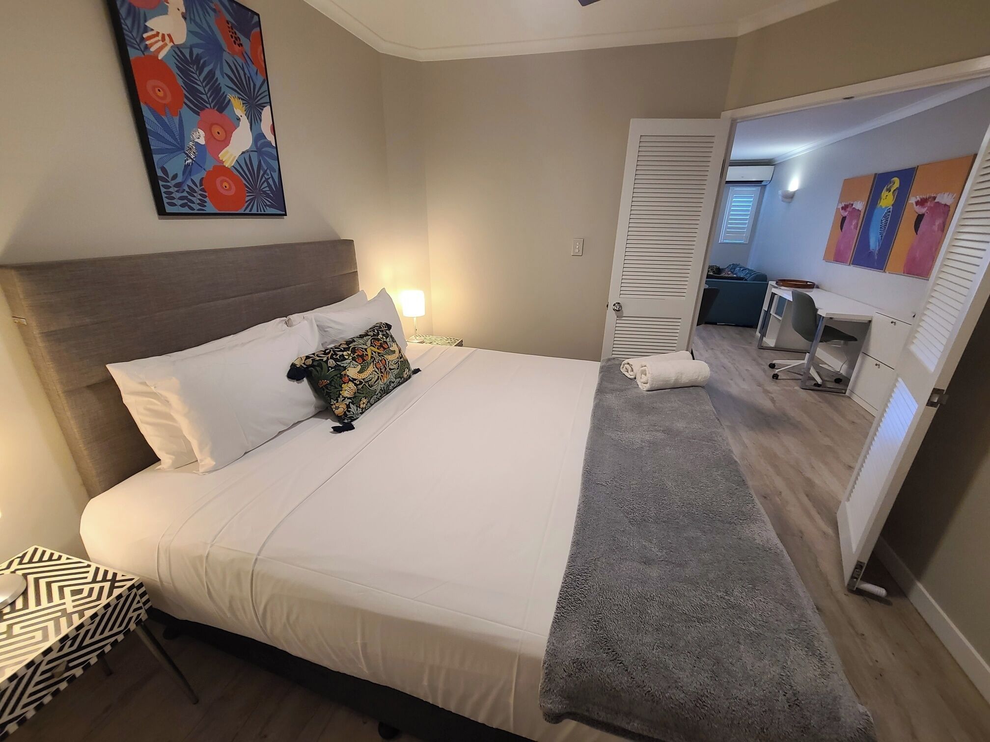 A neatly made bed with white linens and a colorful pillow, a bedside table with a patterned tray, and a door leading to a bathroom with a towel and a mirror.