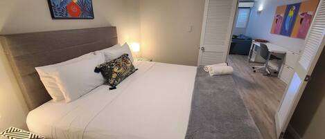 A neatly made bed with white linens and a colorful pillow, a bedside table with a patterned tray, and a door leading to a bathroom with a towel and a mirror.